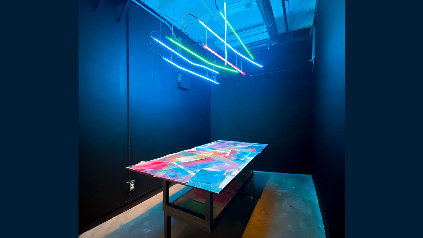 A table with colorful designs underneath glowing sticks hanging from the ceiling in a room with dark blue walls.