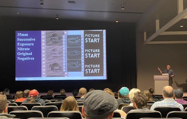 Facing a stage towards the back of an auditorium, Chase Schulte stands at a podium on the right facing the audience, with a large PowerPoint slide projected in the middle of the room. The left of the slide reads "35mm Successive Exposure Nitrate Original Negatives." The right of the slide shows three examples of nitrate film reel.