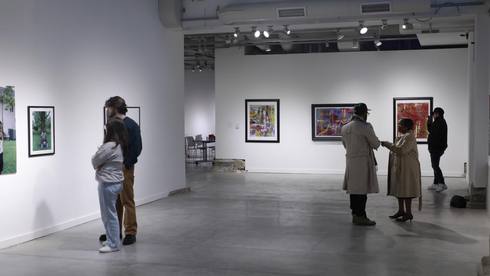 In the gallery, three people are admiring artwork and two other people are in conversation.